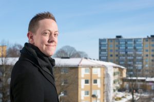 Håkan Eriksson står snett mot kameran och ler. I bakgrunden syns höghus.