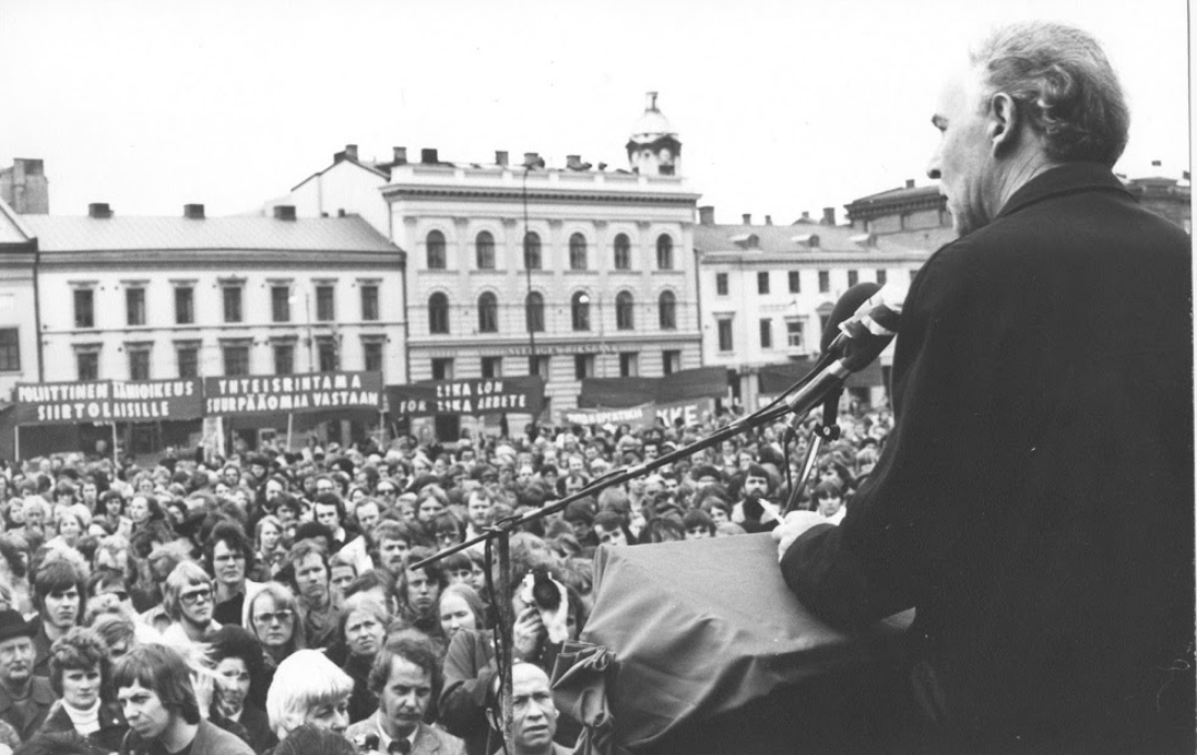 Svartvit bild tagen bakifrån på C.-H. Hermansson, Vänsterpartiets dåvarande ordförande, håller tal på första maj någon gång på 70-talet. I bakgrunden syns den stora åhörarskaran.