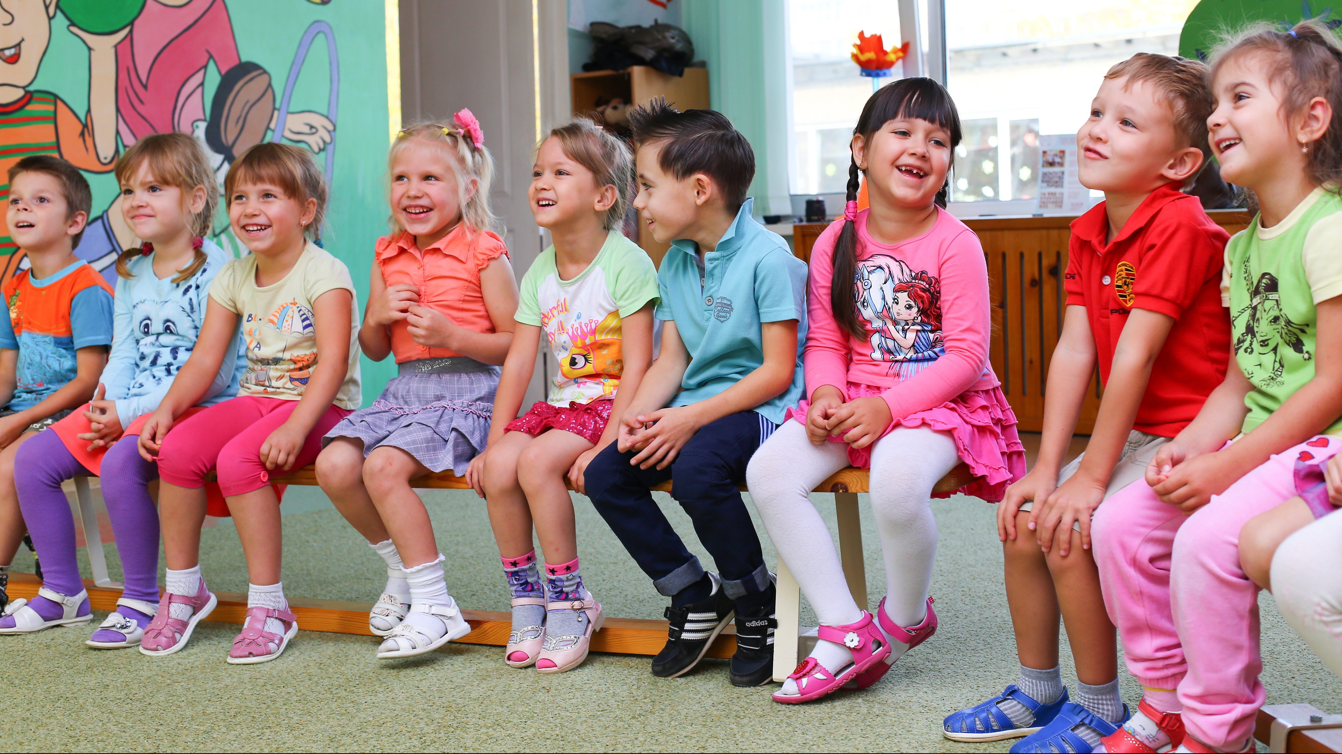 Tio leende barn i färgglada kläder sitter på en bänk i en halvcirkel. Bakom barnen syns ett fönster och en öppen dörr syns till vänster. Väggen på vänster sida är målad i ljust grön färg med två lekande barn. Golvet är klätt med ljusgrön heltäckningsmatta.