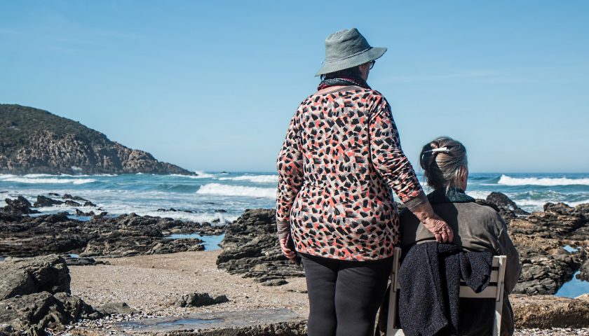 TVå älder kvinnor blickar ut över havet. De har ryggen mot betraktaren. En sitter i en solstol.